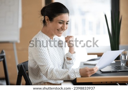 Overjoyed young indian woman employee sit at desk read good news in paper post correspondence. Excited ethnic female worker feel euphoric triumph get pleasant message in paperwork document. Royalty-Free Stock Photo #2398833937