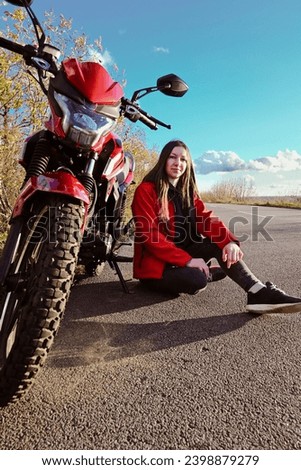 young attractive biker girl in red sitting next to her red motorcycle in the middle of the road. biker with a motorcycle outside the city.