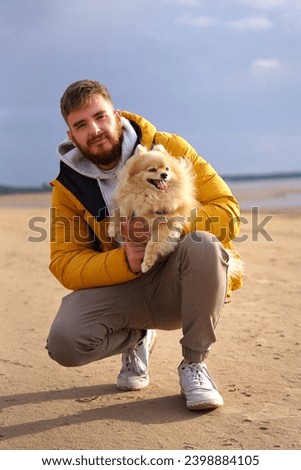 Happy young man walking with his pet Pomeranian Spitz dog on the beach, hold puppy on hands. People love animals concept. Vertical photo.