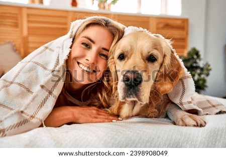 Similarity between dog and owner. Portrait of cheerful woman with blond hair snuggling to adorable golden retriever while lying together on comfy bed under soft blanket. Royalty-Free Stock Photo #2398908049