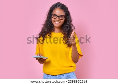 Young cheerful ethnic Indian woman teen with mobile phone in hands points finger at camera and looks at screen inviting to chat on social network or instant messenger stands posing in pink studio. Royalty-Free Stock Photo #2398932369