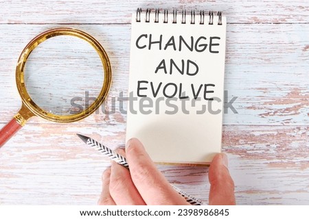 CHANGE AND EVOLVE written on a blank sheet of a notebook which is touched by a man's hand with a pencil and a magnifying glass is lying next to it Royalty-Free Stock Photo #2398986845