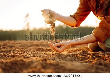 Young female farmer's hands touch dry soil in an agricultural field. A woman agronomist sorts through the black soil, checking the quality of the soil before sowing. Gardening and ecology concept. Royalty-Free Stock Photo #2399015201