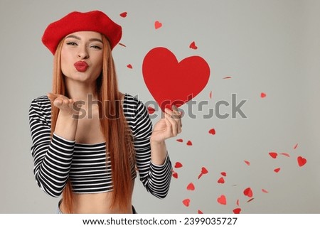Young woman with paper heart blowing kiss under confetti on light grey background, space for text