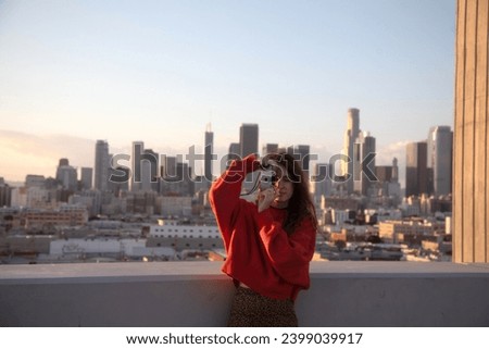 A young beautiful brunette girl with an old film photo camera in front of LA downtown panoramic view, taking a photo.