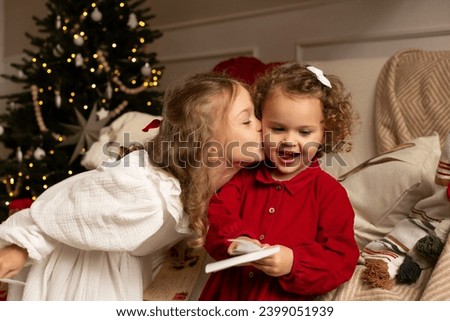 Two little girls kissing while writing the letter to Santa Claus near Christmas tree indoors. Merry Christmas and Happy Holidays.