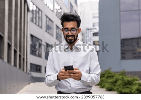 Young successful businessman walking down the street holding a phone in his hands, an Indian man using an application on a smartphone smiling contentedly from outside an office building.
