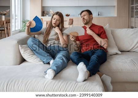 Unhappy couple sitting on sofa waving with hand fans because of broken conditioner, absence of ventilator, air blower. Wife and husband feeling unwell, dehydrated, having difficult breath, headache Royalty-Free Stock Photo #2399140309