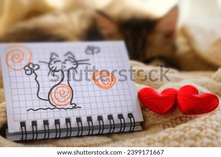 Fun picture on paper, notepad and two red hearts. Pretty cute kitten relaxing on the bed. Tabby cat. Funny pets.