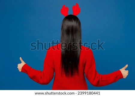 Back rear view merry young Latin woman wear red Christmas sweater decorative fun deer horns on head posing show thumb up like gesture isolated on plain blue background. Happy New Year holiday concept