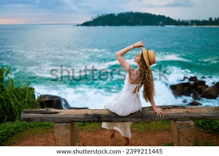 Young woman in a flowing white sundress and straw hat sits on a rustic wooden bench by the sea, contemplating the waves at dusk, embodying peace and solitude. Royalty-Free Stock Photo #2399211445