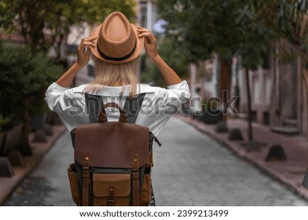 Stylish young woman in a white shirt, skirt with a craft backpack holds her hat with her hands while standing in the middle of a beautiful street, Turkey. Concept of travel, tourism. Vertical photo