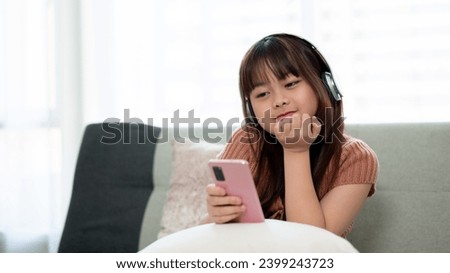 Young adorable Asian girl in casual clothes wearing headphones and using her smartphone, enjoying listening to music on a sofa in the living room. Kid and technology concepts