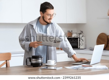Young man with newspaper using laptop in kitchen