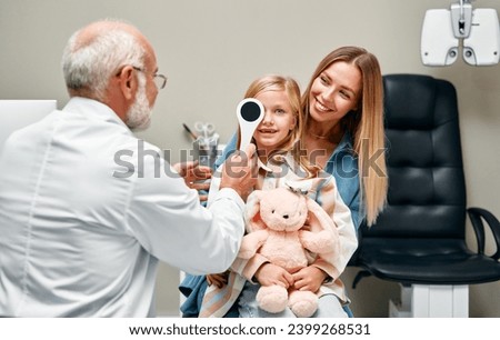 Ophthalmologist closing one eye of a cute little girl while sitting on her mother's lap during a consultation. A child points to an eye chart during an eye test. Royalty-Free Stock Photo #2399268531