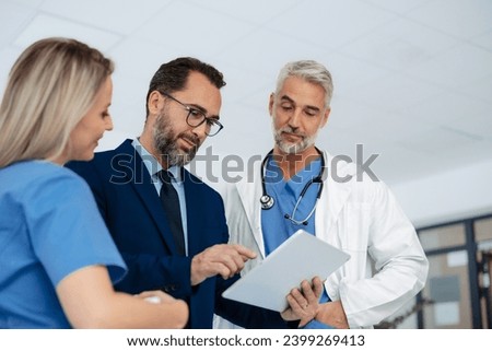 Pharmaceutical sales representative talking with doctors in medical building, presenting them new product on tablet, business talk. Hospital director consulting with healthcare staff. Royalty-Free Stock Photo #2399269413