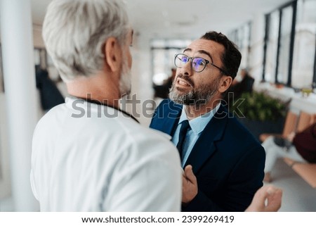 Violence against health workers. Patient is aggressive, have conflict with doctor in hospital corridor. Visitor holding doctor by coat, screaming, threatening him. Royalty-Free Stock Photo #2399269419