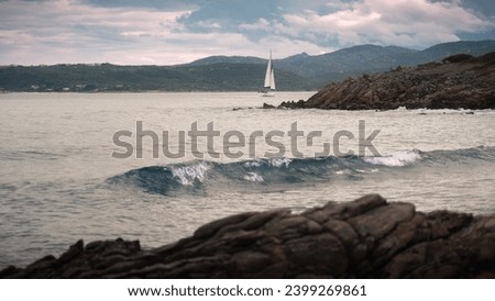 In the distance a sailing boat with its sails unfurled sails the sea in a winter Mediterranean landscape, a wave in the foreground dampens the calm of the sea.