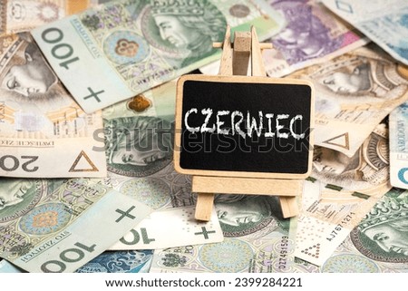 a small wooden writing board standing on scattered Polish zloty PLN banknotes, a chalk inscription "Czerwiec" on the black board. translation: June (selective focus)