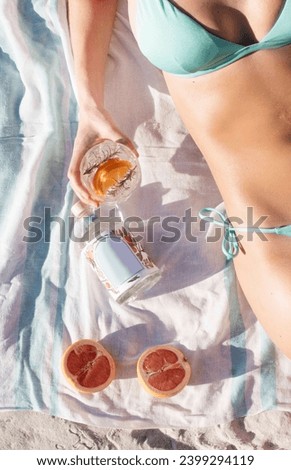 Flat lay of aucasian woman lying on linen beach towel wearing a turcoise bikini. Half of body can be seen and some beach sand. She is holding a clear cocktail with slices of grapefruit. 