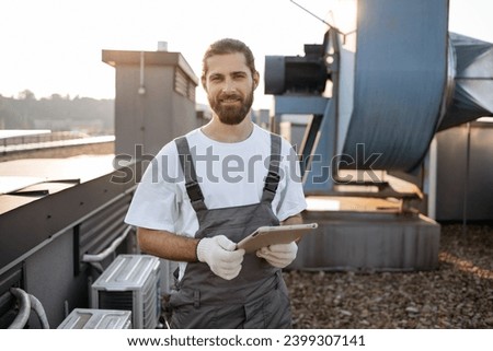 Serious construction worker in overalls and gloves standing and using modern tablet outdoors. Caucasian professional man surfing internet while working with devices on roof of factory. Royalty-Free Stock Photo #2399307141