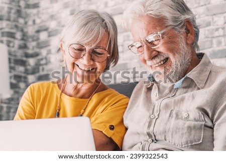 Pretty elderly 70s grey-haired couple resting on couch in living room hold on lap laptop watching movie smiling enjoy free time, older generation and modern wireless technology advanced users concept Royalty-Free Stock Photo #2399322543