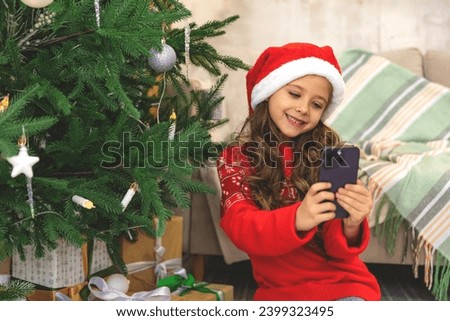 Cute little girl taking a selfie photo by using a smartphone near a Christmas tree. little girl making photo on mobile phone near Christmas tree. Garland