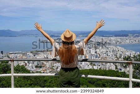 Tourism in Florianopolis, Brazil. Tourist woman with raising arms from belvedere in Florianopolis city, Santa Catarina, Brazil. Royalty-Free Stock Photo #2399324849