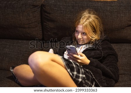 a teenage girl of light appearance sits on a dark sofa, holding a phone in her hands