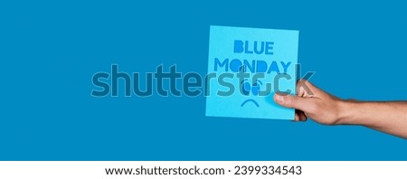 a man holding a blue sign with the text blue monday and a sad face on a blue background with some blank space on the left, in a panoramic format to use as web banner or header Royalty-Free Stock Photo #2399334543