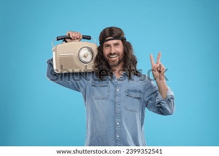 Stylish hippie man with retro radio receiver showing V-sign on light blue background