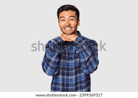 Young Chinese man in studio background keeps hands under chin, is looking happily aside.