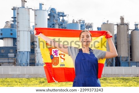 Against backdrop of factory located outside city,balanced girl in overalls of worker stands and holds flag of Spain in her hands.