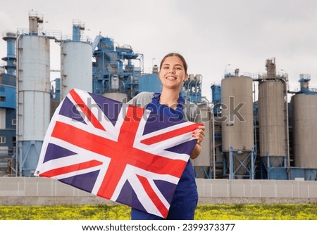 Happy young woman worker holding big flag of Great Britain against background of factory