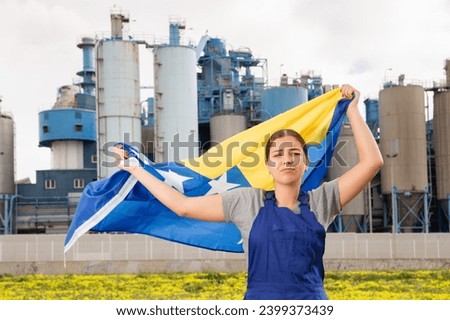 in countryside,worker in overalls,frustrated with working conditions,stands in front of factory building and holds Bosnian flag in her hands Royalty-Free Stock Photo #2399373439