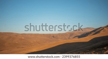 Sunset light casts a warm glow on the smooth, undulating contours of a grass-covered highland, with shadowy mountain ridges in the background under a soft sky Royalty-Free Stock Photo #2399376855