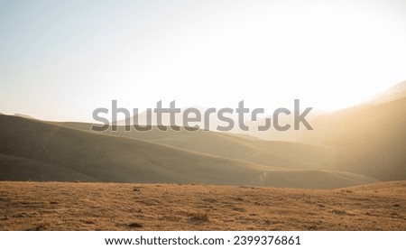 Soft sunlight filters over undulating grassy hills, casting a warm glow on the serene highland landscape with distant mountain silhouettes. Royalty-Free Stock Photo #2399376861