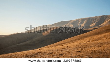 Sunset light casts a warm glow on the smooth, undulating contours of a grass-covered highland, with shadowy mountain ridges in the background under a soft sky Royalty-Free Stock Photo #2399376869