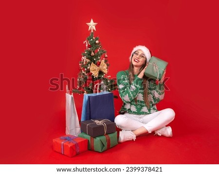 Shaking gift box, full body view young Santa woman sit near decorated Christmas tree shaking gift box. Wondering what's in inside present. Winter holiday, happy new year celebration concept idea.