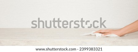 Young adult woman hand holding white dry paper napkin and wiping beige stone tabletop at home kitchen. Closeup. Wide banner. Cleaning service. Front view. Empty place for text on wallpaper background. Royalty-Free Stock Photo #2399383321
