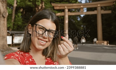 Cheerful young hispanic woman in glasses captures her heart-shaped hand sign in a selfie at japan's iconic meiji temple, exuding love and happiness in this beautiful picture