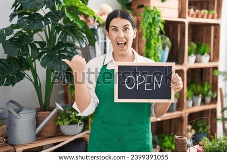 Young hispanic woman working at florist holding open sign pointing thumb up to the side smiling happy with open mouth 