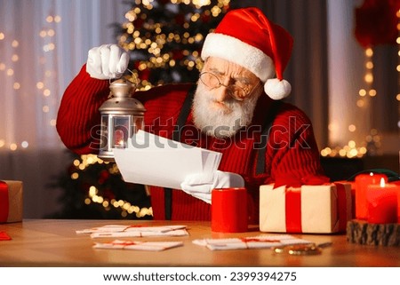 Santa Claus holding lantern and reading letters at his workplace in room decorated for Christmas