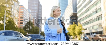 Street stylish portrait of young happy woman, blond girl with backpack and laptop, standing outdoor s near road on sunny bright day. Lifestyle concept