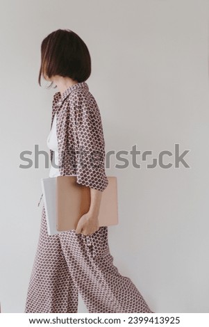 Laptop computer and paper documents in female hand. Young pretty woman in pajamas stays over white wall. Businesswoman, work at home concept Royalty-Free Stock Photo #2399413925