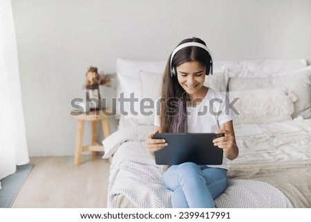 Attractive pre-teen 12s girl wear wireless headphones using modern digital tablet seated on bed in cozy bedroom. I generation and technologies usage for fun, education and leisure