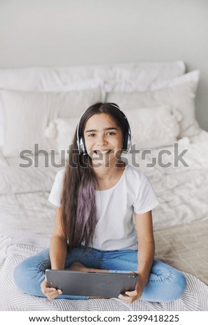 Attractive pre-teen 12s girl wear wireless headphones using modern digital tablet seated on bed in cozy bedroom. I generation and technologies usage for fun, education and leisure