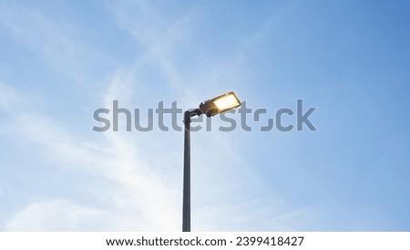 Location photos Tall black light pole on the side of the road Alone in the middle of the picture Has a warm yellow light. The background is faint clouds under the evening blue sky. The atmosphere