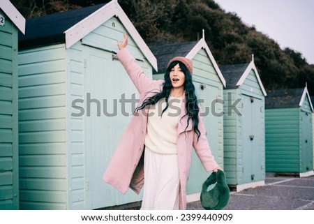 Stylish emotional hipster woman with color hair wearing pink coat, hat and fur bag having fun on mint green beach hut background. Seasonal city street fashion. Barbiecore style. Selective focus. Royalty-Free Stock Photo #2399440909