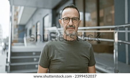 Confident middle age man with grey hair, sporting glasses and a beard, stands at an urban street corner, his serious expression betraying the weight of his thoughts. Royalty-Free Stock Photo #2399452037